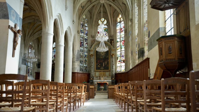 Intérieur de l'Église Saint-Éloi. Nous sommes situés au centre de l'allée principale avec de chaque côté des rangées de chaises. Sur les murs du fond, on voit des vitraux